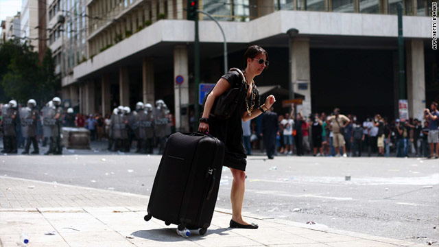 A tourist walks down the sidewalk while protesters line the streets and police stand guard in Greece, on Tuesday.